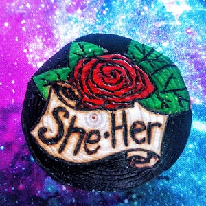 May include: A wooden round pin with a black background, a red rose with green leaves, and the text "She/Her" on a white scroll.