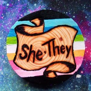 Könnte beinhalten: Eine runde Holzscheibe mit einer bemalten Regenbogenflagge und einer Schriftrolle mit den Worten "She/They", die in das Holz gebrannt sind.