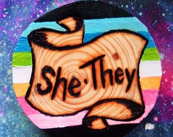 Può includere: Un disco rotondo di legno con una bandiera arcobaleno dipinta e un rotolo con le parole "She/They" bruciate nel legno.