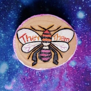 Op de afbeelding: Een rond houten ornament met een roze en zwarte bij erop geschilderd. De bij heeft rode en oranje strepen op zijn achterlijf. De woorden "They/Them" staan op de vleugels van de bij.