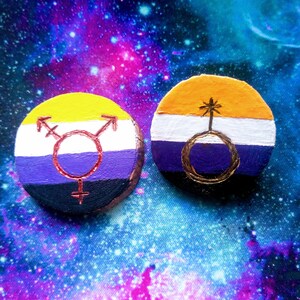 Puede incluir: Dos adornos redondos de madera pintados con los colores de las banderas del orgullo genderqueer y no binario. La bandera genderqueer tiene rayas amarillas, moradas y negras con un símbolo transgénero dorado. La bandera no binaria tiene rayas amarillas, blancas, moradas y negras con un símbolo dorado de un círculo con una estrella dentro.