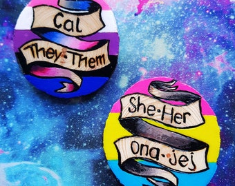 Può includere: Due ornamenti rotondi in legno dipinti con i colori delle bandiere del pride pansexuale e transgender. L'ornamento pansexuale ha un motivo a strisce gialle, blu e rosa con un nastro nero che dice "She-Her Ona-Jej". L'ornamento transgender ha un motivo a strisce blu chiaro, rosa e bianco con un nastro nero che dice "Cal They-Them".