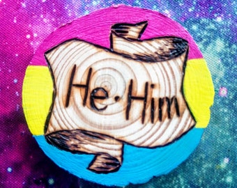 Può includere: Un cerchio di legno dipinto con i colori della bandiera del pride pansexuale, rosa, giallo e blu. Il cerchio ha un design in legno bruciato di uno scroll con le parole "He/Him" scritte su di esso.