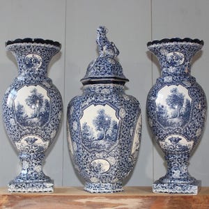 Può includere: Tre vasi in ceramica blu e bianca con un disegno a paesaggio. I vasi presentano un motivo floreale e geometrico dettagliato. Il vaso centrale ha un coperchio con una piccola statuina di animale. Vasi decorativi.