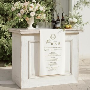 Luxury wedding bar sign on ivory linen fabric, styled with greenery and glassware on rustic wooden bar table