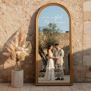 Puede incluir: Un gran espejo dorado arqueado con el texto "WE'RE SO glad YOU ARE HERE" y nombres y fecha. El espejo refleja a una pareja tomados de la mano. Un arreglo floral seco está a la izquierda.
