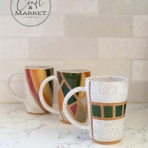 May include: Three ceramic mugs with unique designs. One mug features a white speckled finish with green and brown geometric shapes. Another has a colorful striped pattern, and the third has a geometric design with green, orange, and brown shapes. The mugs have large handles.
