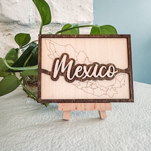 May include: A wooden sign featuring a map of Mexico with the country's name in a cursive font. The sign is displayed on a small wooden easel. The sign has a light wood background with a dark brown border and lettering.