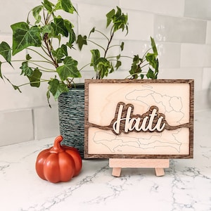 May include: Wooden Haiti map art with a light wood finish and a dark wood frame. The word "Haiti" is cut out in a cursive font and layered on top. A small orange pumpkin and a potted plant are in the background.