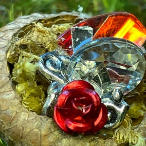 May include: A detailed shot of a decorative piece featuring a red rose, clear and red crystals, and silver embellishments nestled within a pine cone. The background is blurred, with green foliage.