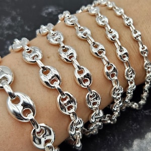 May include: A set of silver mariner chain bracelets of varying widths. The bracelets are stacked on a person's wrist.