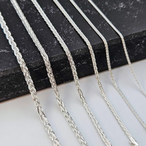 Puede incluir: Un conjunto de siete cadenas de plata de diferentes anchos, mostradas sobre una superficie negra. Las cadenas son todas de diseño trenzado.