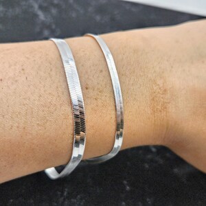 May include: Two silver snake chain bracelets stacked on a person's wrist.