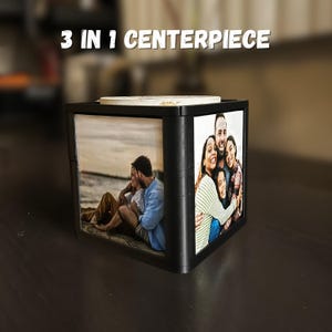 May include: Black cube-shaped centerpiece with family photos on three sides. The top has a light-colored surface. The text "3 IN 1 CENTERPIECE" is displayed above the cube. The photos show a couple on a beach and a family hugging.