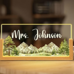 May include: A rectangular wooden-framed LED light with the name "Mrs. Johnson" in white script. The illuminated panel features a watercolor landscape of mountains and pine trees. The light is on a wooden table.