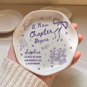 May include: A white ceramic dish with a gold rim, featuring purple text and illustrations. The text reads "a New Chapter Begins" and includes the name "Sophia", "Class of 2026", and "University of California".
