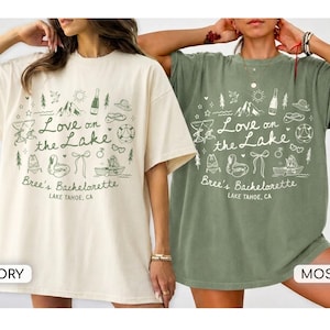 May include: Two oversized t-shirts in ivory and moss green featuring a dark green graphic print. The design includes the text "Love on the Lake", "Bree's Bachelorette", and "LAKE TAHOE, CA" surrounded by illustrations of mountains, a boat, and drinks.