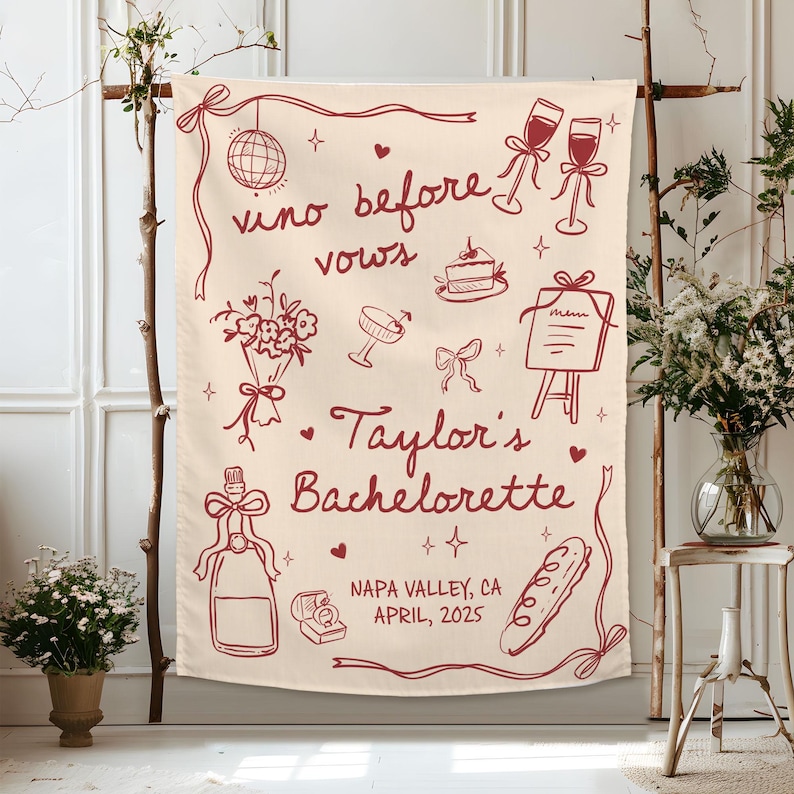 Op de afbeelding: Een cr&egrave;mekleurige stoffen banner met donkerrode lijntekeningen van een discobal, champagneglazen, een taart, een boeket, een menukaart, een fles en een ringdoosje. De tekst luidt: vino before vows, Taylor's Bachelorette, NAPA VALLEY, CA, APRIL, 2025.