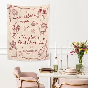 Op de afbeelding: Een cr&egrave;mekleurige stoffen wandhanger met donkerrode lijntekeningen van een discobal, champagneglazen, taart, bloemen, een menu, een fles en een stokbrood. De tekst luidt: "vino before vows", "Taylor's Bachelorette" en "NAPA VALLEY, CA APRIL, 2025".