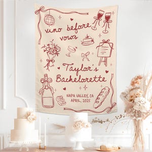 Op de afbeelding: Een cr&egrave;mekleurige stoffen wandhanger met donkerrode lijntekeningen van een discobal, wijnglazen, een boeket, een taart, een menu, een fles, een ringdoosje en een stokbrood met de tekst "vino before vows", "Taylor's Bachelorette" en "NAPA VALLEY, CA APRIL, 2025".