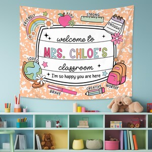 May include: A peach-colored classroom tapestry with a white border and a welcome sign that reads "Welcome to Mrs. Chloe's Classroom". The sign is surrounded by colorful illustrations of school supplies, including a rainbow, a globe, a pencil, a ruler, a backpack, and a notebook. The tapestry also features the words "Friendly", "Kind", "Strong", "Enough", "Loved", "Brave", "Creative", and "Worthy" in a colorful font.