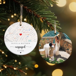 May include: Two round Christmas ornaments hanging from a pine branch. One ornament features a map with a red heart and the text "Olivia & Matthew engaged 20.10.2025". The other ornament shows a couple during a proposal.