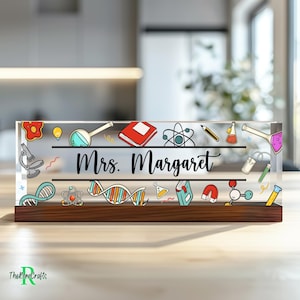 May include: A clear acrylic desk nameplate with a wooden base. The nameplate features a colourful design of science-related icons and the text "Mrs. Margaret".