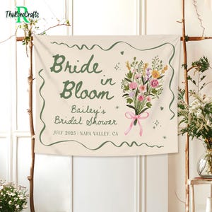 May include: A cream-colored fabric banner featuring the dark green text Bride in Bloom, Bailey's Bridal Shower, and JULY 2025 | NAPA VALLEY, CA. A watercolor illustration of a colorful wildflower bouquet tied with a pink ribbon is on the right.