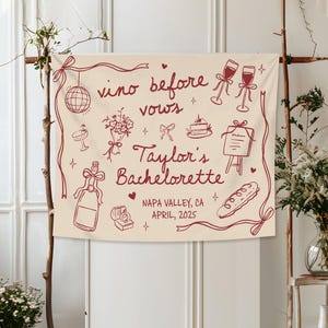 Op de afbeelding: Een cr&egrave;mekleurige stoffen banner met donkerrode lijntekeningen van een discobal, wijnglazen, een boeket, een taart, een menu, een fles, een ringdoosje en een stokbrood. Tekst: vino before vows, Taylor's Bachelorette, NAPA VALLEY, CA, APRIL, 2025.