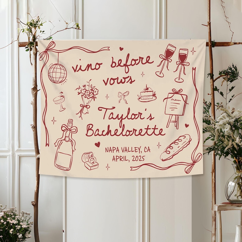 Op de afbeelding: Een cr&egrave;mekleurige stoffen banner met donkerrode lijntekeningen van een discobal, wijnglazen, een boeket, een taart, een menu, een fles, een ringdoosje en een stokbrood. Tekst: vino before vows, Taylor's Bachelorette, NAPA VALLEY, CA, APRIL, 2025.