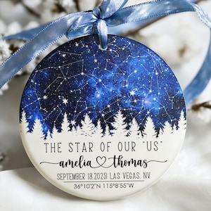 May include: A circular ceramic ornament with a blue and white starry night design. The ornament has the text "THE STAR OF OUR 'US'" and personalized names and a date. A blue ribbon is attached for hanging.
