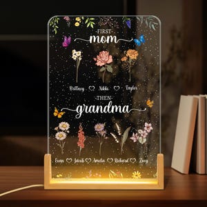 May include: A white illuminated acrylic sign with a floral design and the text "First Mom - Then Grandma" with names listed below. The sign is on a wooden base.