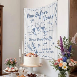 Puede incluir: Una pancarta de tela blanca con rayas verticales azules y texto azul que dice Vino Before Vows, Olivia's Bachelorette, y Nashville, TN June 18-21st, 2026. El dise&ntilde;o presenta ilustraciones de copas de vino, una botella y lazos.