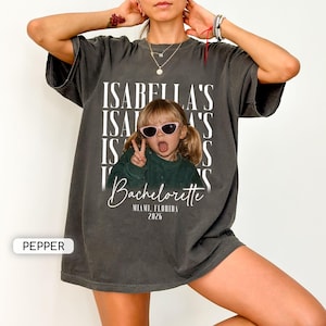 May include: A charcoal gray oversized t-shirt featuring a graphic print of a child wearing pink sunglasses and making a peace sign. The text reads ISABELLA'S, Bachelorette, MIAMI, FLORIDA, 2026. The shirt has a crew neckline and short sleeves.