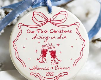 May include: A white ceramic Christmas ornament with red text and illustrations. The ornament features the text "Our First Christmas Living in Sin", two champagne glasses, and the names "Thomas & Emma" with the year "2025". A blue ribbon is attached.