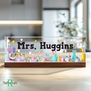 May include: A clear acrylic desk nameplate with a wooden base. The nameplate features a miniature kitchen scene with colourful kitchenware and the text "Mrs. Huggins".