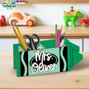 以下が含まれることがあります： 緑と黒のクレヨン型のデスクオーガナイザー。白い筆記体で「Mrs. Oliver」と書かれています。鉛筆、ペン、ハサミが入っています。背景にはおもちゃの車と緑のカバがあります。