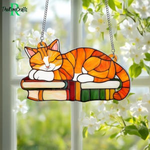 May include: A stained glass window hanging features an orange tabby cat sleeping on a stack of colorful books. The cat has white paws and a white patch on its chest. The books are red, green, yellow, and blue.