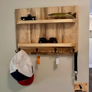 May include: A wooden wall-mounted shelf with two shelves and five hooks. The top shelf has a toy truck and a fish figurine. The bottom shelf has a pair of sunglasses. The hooks have a hat, keys, and a lanyard hanging from them. The shelf is made of light wood with a natural finish. The text "BLACK DOG WOODWORKS" is visible in the bottom right corner.