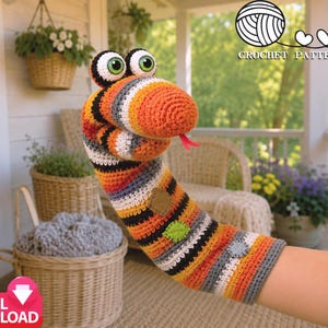 Puede incluir: Una marioneta de serpiente tejida a ganchillo con rayas horizontales en naranja, negro, blanco y gris. La serpiente tiene ojos grandes blancos con pupilas verdes y una pequeña lengua roja. El texto arriba a la derecha dice CROCHET PATTERN PDF.