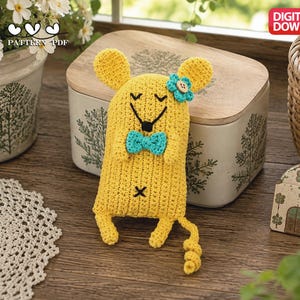 May include: A yellow crocheted mouse toy featuring a teal bow tie, a teal flower with a wooden button center on its head, and embroidered black eyes and nose. The mouse has a curly tail and a small black cross stitch on its lower body. Digital download.