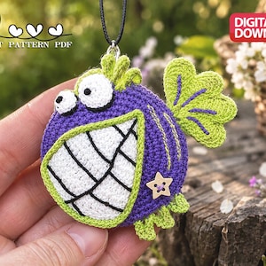 May include: A purple and lime green crocheted fish ornament featuring large white eyes, a wide white mouth with black stitched teeth, a wooden star-shaped button, and a black cord for hanging. The text CROCHET PATTERN PDF is visible.