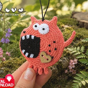 May include: A coral-colored crochet monster ornament featuring two white eyes with black pupils, a large black mouth with white teeth, and a wooden heart-shaped button. The creature has small legs and a tail, hanging from a black string. Text reads CROCHET PATTERN PDF.
