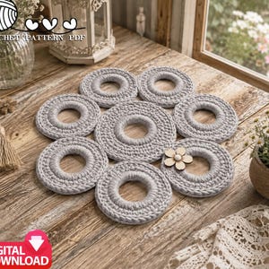 May include: A crochet pattern PDF featuring a light gray, circular, ring-shaped mat composed of seven interconnected, crocheted rings arranged in a flower-like pattern, with a small, light-colored wooden flower embellishment on one ring.