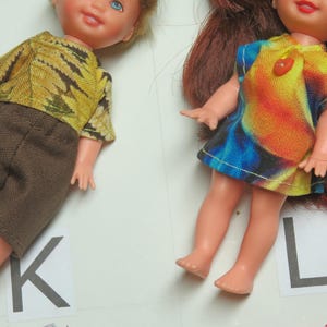 May include: Two dolls are shown. One doll wears a yellow and brown patterned top with brown shorts. The other doll wears a colorful tie-dye dress with a red heart-shaped button. The dolls are on a white surface.