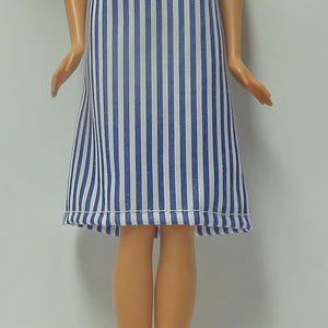 May include: A doll wearing a blue and white striped dress. The dress has a simple, sleeveless design with vertical stripes and a ruffled hem. The doll has blonde hair and is standing against a white background.