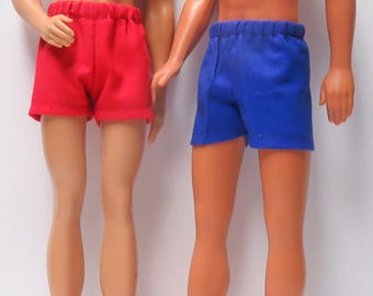 May include: Two male dolls are shown, one in red shorts and the other in blue shorts. The dolls are standing close together, with their arms touching. The shorts are simple, elastic-waisted designs.