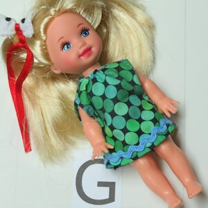 May include: A small doll with blonde hair, blue eyes, and pink lips, wearing a sleeveless green and black polka dot dress with a light blue wavy trim at the hem. A white butterfly hair accessory with a red ribbon is attached to the doll's hair.