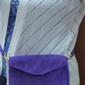 May include: A white knitted cardigan with a purple suede crossbody bag. The bag has a flap closure and a gold chain strap.