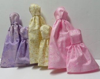 Peut inclure: Quatre robes de poupée de différentes tailles et couleurs. Les robes sont en tissu scintillant avec un motif subtil. Les couleurs sont lavande, or, rose et rose clair.